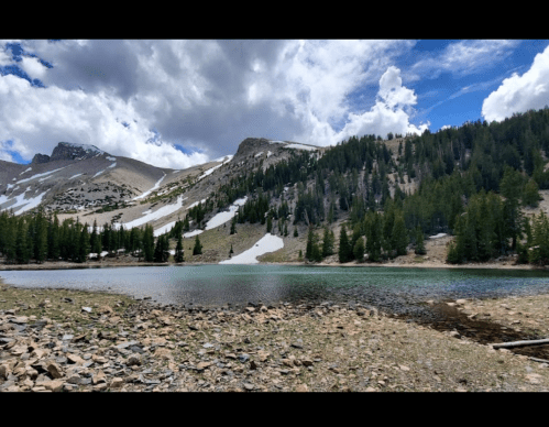 A serene mountain landscape featuring a clear lake surrounded by trees and rocky terrain under a partly cloudy sky.