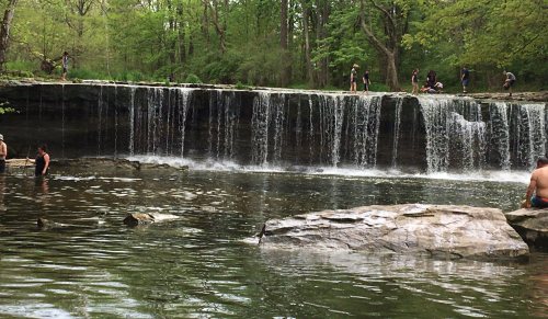 A serene waterfall cascades into a calm pool, surrounded by lush greenery and people enjoying the natural setting.