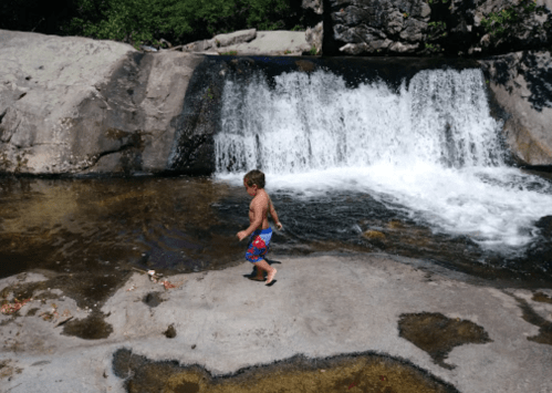 A young boy in swim trunks walks near a waterfall, splashing in the shallow water on a sunny day.