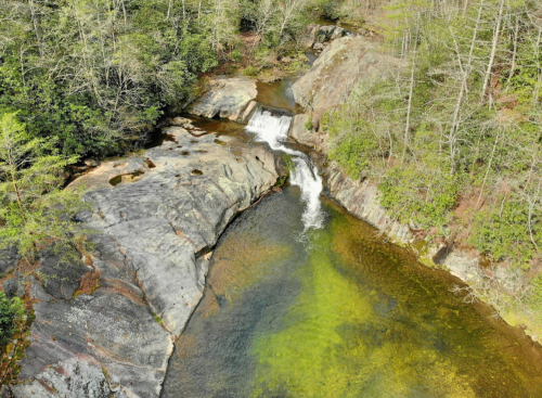 A serene river flows over rocks, creating a small waterfall surrounded by lush greenery and trees.