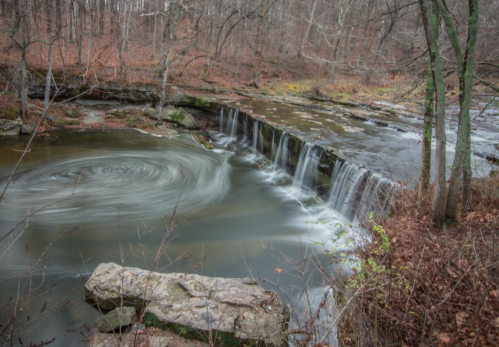 A serene waterfall cascades over rocks, surrounded by autumn foliage and a swirling pool of water.