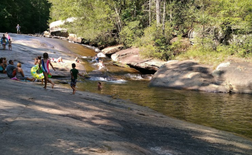 A group of people enjoying a sunny day by a rocky river, with some swimming and others playing on the shore.