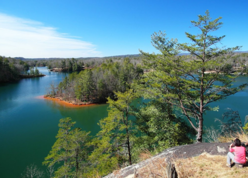 A scenic view of a lake surrounded by trees, with a person sitting on a rock in the foreground.