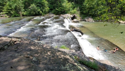 A serene waterfall cascades over rocks into a calm river, with people enjoying the water nearby in a lush green setting.