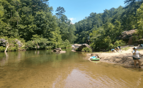 A serene river scene with clear water, sandy banks, and lush green trees, where people enjoy the outdoors.