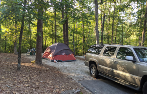 A red and gray tent set up in a wooded campsite, with a parked SUV nearby and trees surrounding the area.