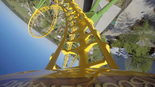 A vibrant yellow roller coaster track spirals upward against a clear blue sky, with green trees in the background.