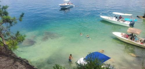 A clear blue lake with boats and people swimming near the shore, surrounded by trees and rocky terrain.