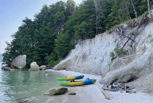Two colorful kayaks on a sandy beach beside a calm water body, with a rocky cliff and trees in the background.