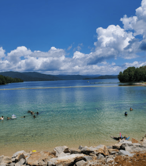 A serene lake with clear blue water, surrounded by trees and mountains, under a partly cloudy sky. People enjoy the water.
