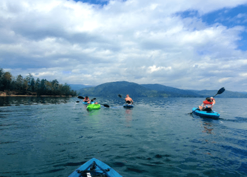 Four people kayaking on a calm lake with mountains and cloudy skies in the background.