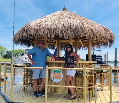 A man and woman pose together at a tiki hut by the water, with boats in the background and a clear blue sky.