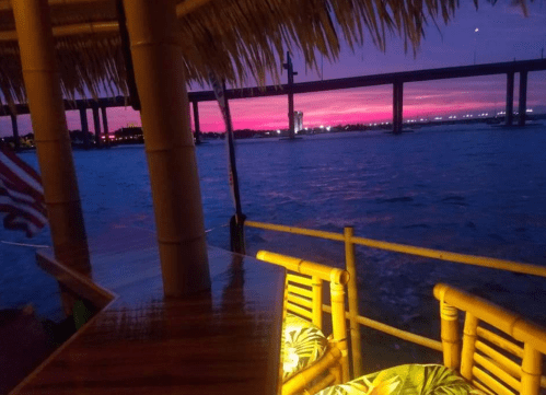 A vibrant sunset over water, viewed from a bamboo structure with a table and chairs, creating a tropical atmosphere.