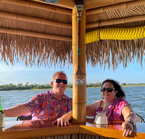 A man and woman sit at a bamboo bar by the water, both smiling and enjoying drinks in a tropical setting.