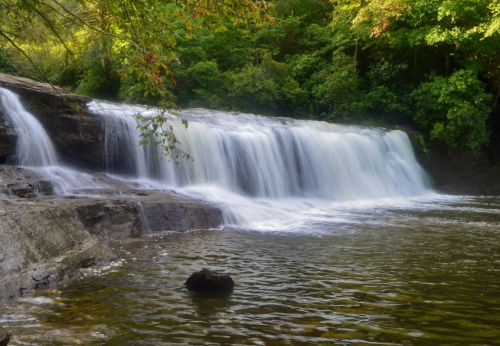A serene waterfall cascades over rocks into a calm pool, surrounded by lush green trees.