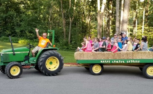 A man waves from a green tractor pulling a hayride filled with smiling children in a wooded area.