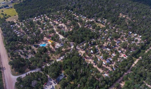 Aerial view of a wooded campground with numerous RVs and tents, surrounded by trees and a swimming pool area.