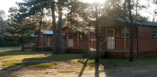 Four wooden cabins with porches, surrounded by trees, illuminated by sunlight in a serene outdoor setting.