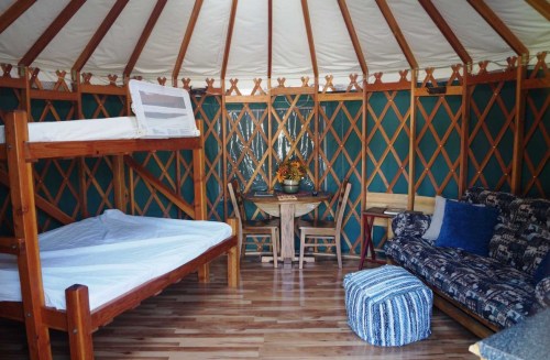 Interior of a yurt featuring a bunk bed, a table with chairs, and a cozy seating area with a patterned couch.