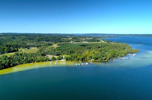 Aerial view of a serene lake surrounded by lush greenery and small boats along the shoreline. Clear blue sky above.