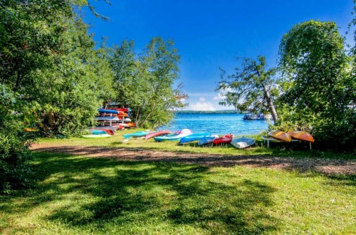 A serene lakeside scene with colorful kayaks on the grass, surrounded by trees and a clear blue sky.