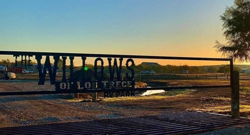 A metal gate with "The Willows of Los Trece RV Park" sign at sunset, with a clear sky and open landscape in the background.