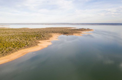 Aerial view of a calm lake with a sandy shore and lush greenery along the water's edge under a clear sky.