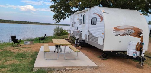 A parked RV by a lake, with a picnic table and two chairs set up nearby under a tree.