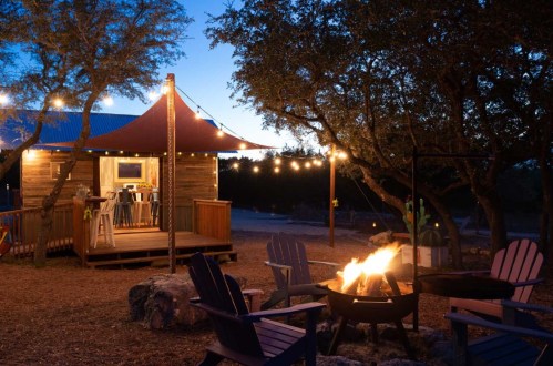 Cozy cabin with string lights at dusk, surrounded by trees and a fire pit with chairs in a serene outdoor setting.