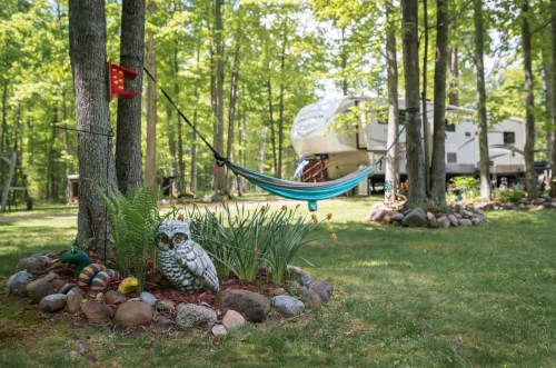 A serene outdoor scene featuring a hammock between trees, a decorative owl, and a camper in a lush green setting.
