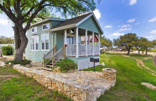 A charming green cottage with a porch, surrounded by trees and a stone pathway, set in a grassy landscape.