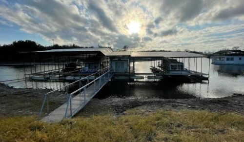 A boat dock with covered slips on a calm lake, under a cloudy sky with the sun peeking through.
