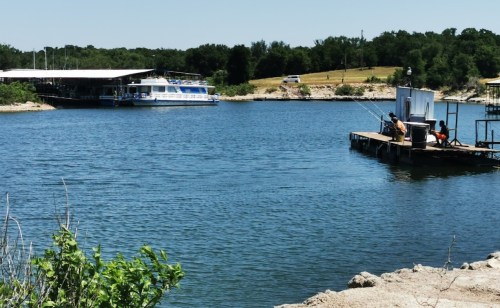 A calm lake scene with a boat dock, fishermen on a pier, and a houseboat in the background under a clear blue sky.