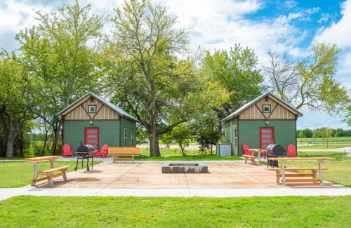 Two green cabins with red doors surrounded by trees, benches, and a fire pit on a sunny day.