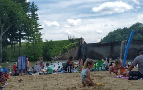 A sandy beach scene with families enjoying the water, a dam in the background, and children playing.