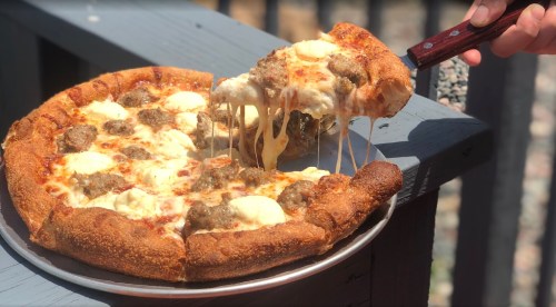 A hand lifts a slice of cheesy pizza topped with sausage, showcasing its gooey cheese and golden crust.
