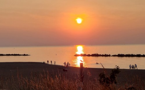 Sunset over a calm lake, with silhouettes of people on the shore and gentle waves reflecting the orange sky.