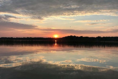 A serene sunset over a calm lake, with vibrant colors reflecting on the water and silhouettes of trees in the background.