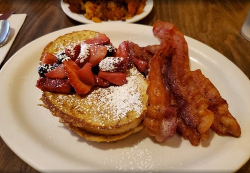 A plate of pancakes topped with strawberries and powdered sugar, accompanied by crispy bacon strips.
