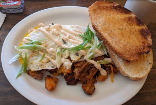 A plate with a fried egg on a bed of potatoes and meat, drizzled with sauce, alongside two slices of toasted bread.