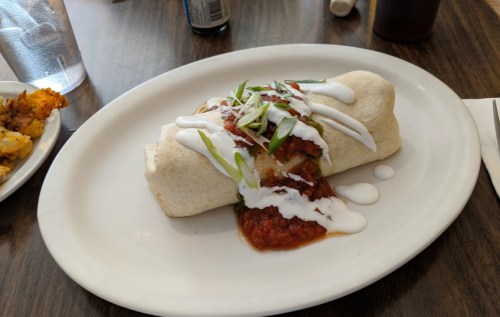 A burrito topped with salsa, sour cream, and green onions on a white plate, with a drink and side dish in the background.
