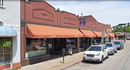 A street view of a brick building with orange awnings, featuring a restaurant and parked cars along the curb.