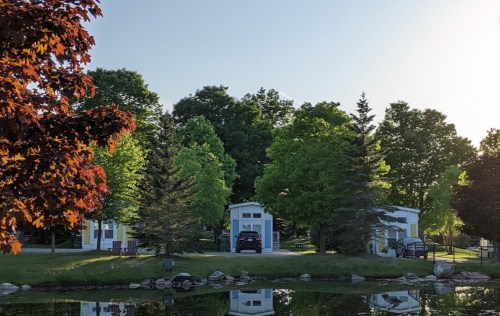Two white buildings surrounded by lush green trees, reflecting in a calm pond under a clear sky.