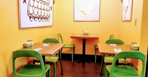Cozy restaurant corner with yellow walls, wooden tables, and green chairs, featuring chicken-themed artwork.