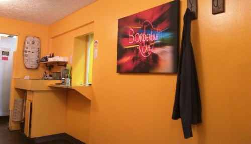 Interior of a cafe with orange walls, a clock, and a colorful "Borderline Cafe" sign. A coat hangs on the wall.