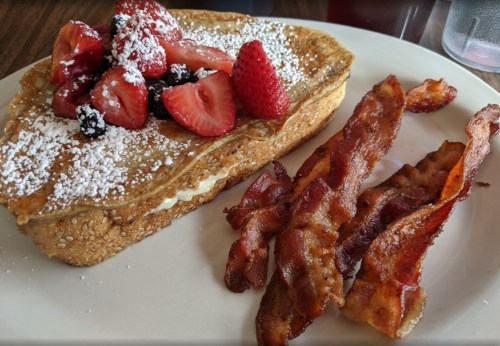 A plate of French toast topped with mixed berries and powdered sugar, served with crispy bacon strips.