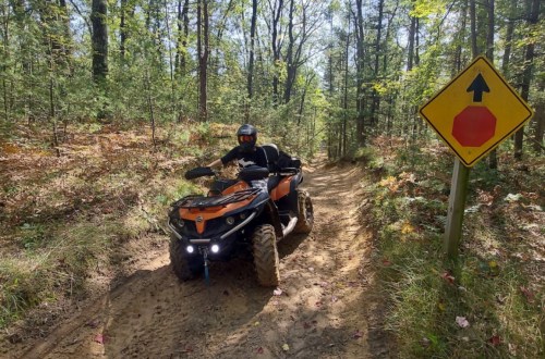 A person rides an orange ATV on a dirt trail surrounded by trees, near a traffic sign indicating a stop ahead.