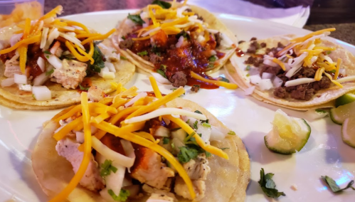 A plate of four tacos topped with chicken, beef, cheese, onions, cilantro, and salsa, with lime wedges on the side.