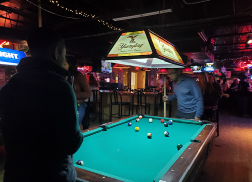 A dimly lit bar with a pool table, neon lights, and people socializing in the background.