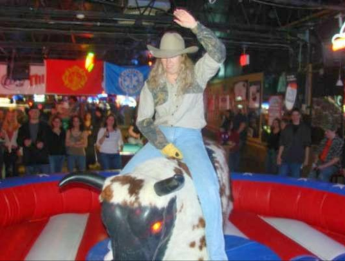 A person in a cowboy hat rides a mechanical bull in a lively bar, surrounded by an enthusiastic crowd.